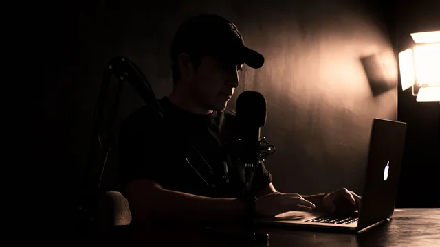 A man wearing a cap works on a laptop with a microphone setup in a dimly lit room. Paid creator memberships